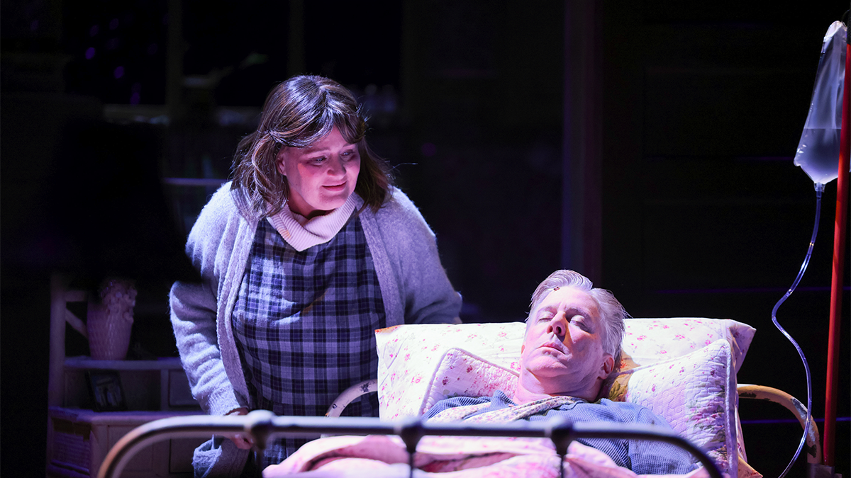 A woman stands over a wounded man in bed. Both on stage.