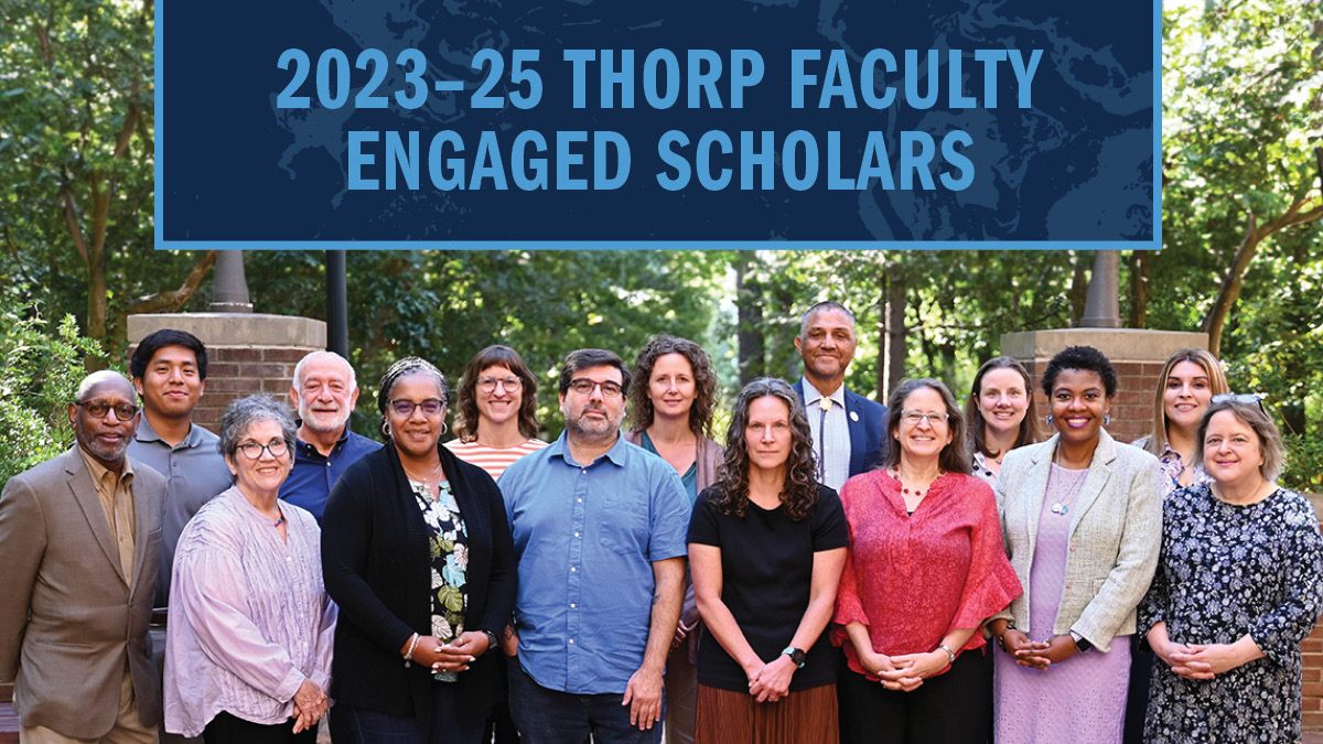A group photo of 15 faculty members standing together and posing for a photo outside in front of a brick wall with trees behind them.