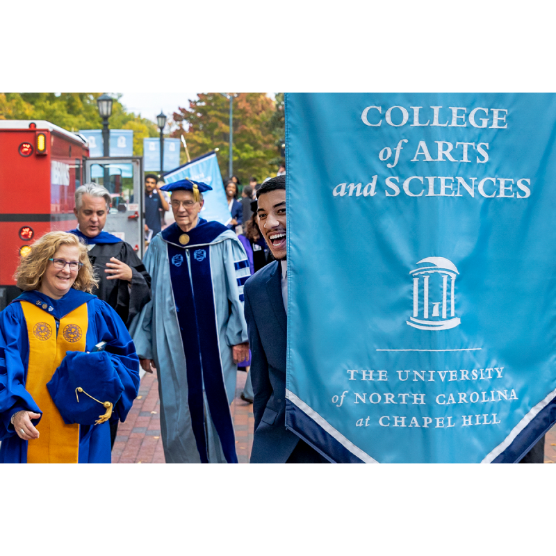 Group of four people, three of whom are in regalia, walking on a brick pathway as part of a University Day ceremony on the campus of UNC-Chapel Hill. A student is holding a banner reading 