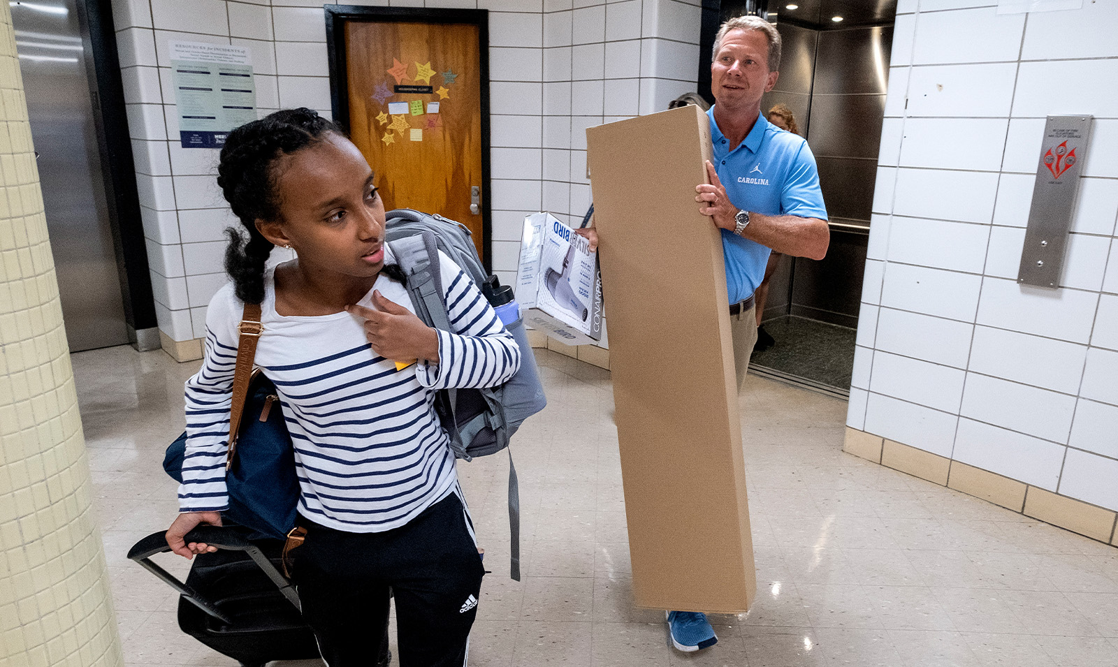 Chancellor Kevin M. Guskiewicz carrying a long, vertical box as he helps a student move into her dormitory on the campus of UNC-Chapel Hill.