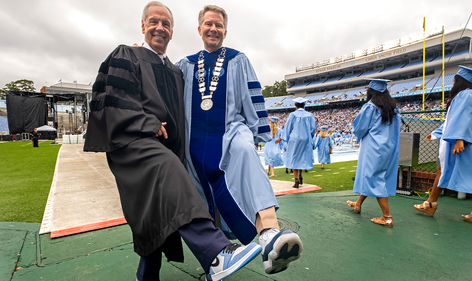 Chancellor Kevin M. Guskiewicz and former Tar Heels basketball coach Roy Williams in regalia at a graduation ceremony showing off their basketball shoes.