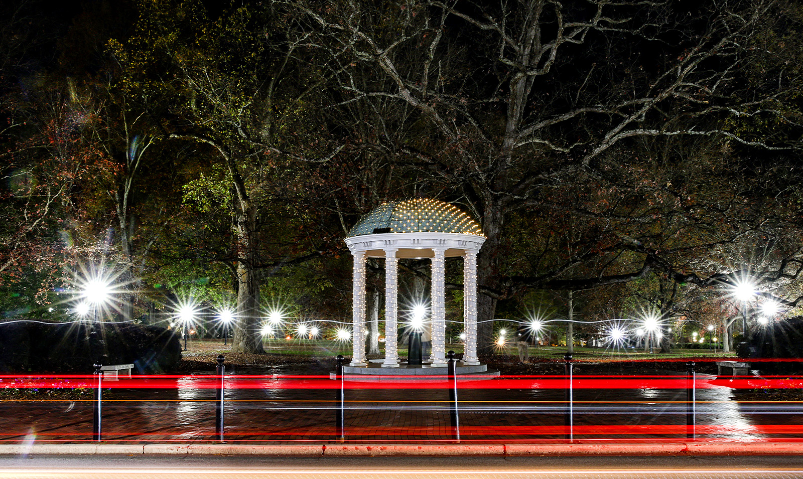 Nightime view of the Old Well on the campus of UNC-Chapel Hill. Long-exposure car lights of passing-by cars on Cameron Avenue are seen in the foreground, and trees and lights from McCorkle Place are seen behind the well.