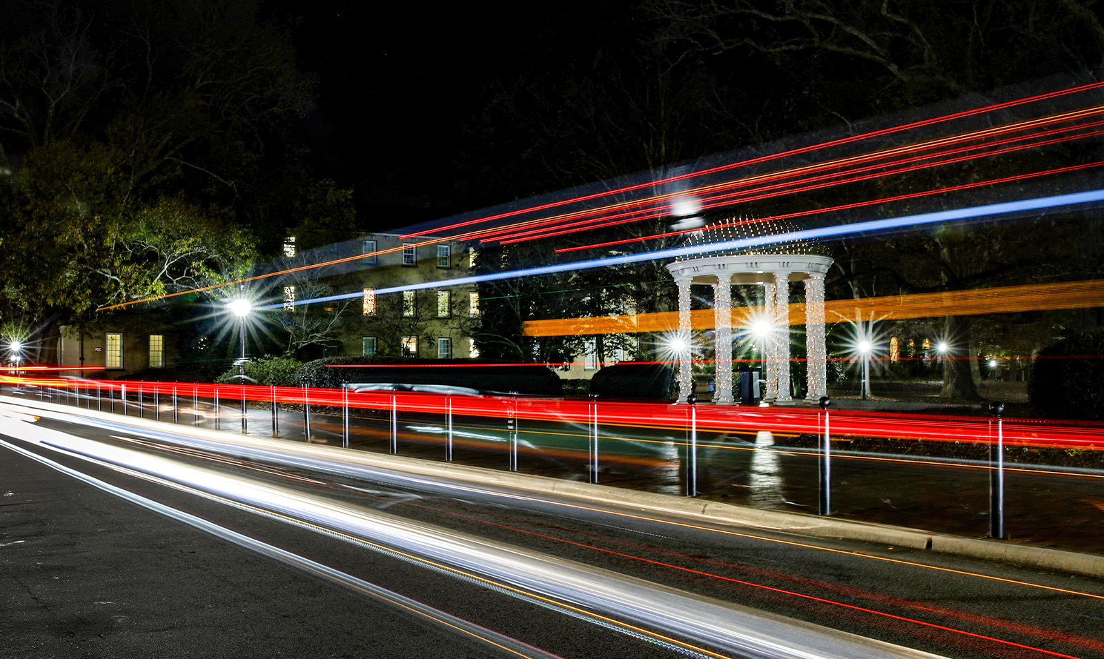 Nightime view of the Old Well decorated with holiday lights on the campus of UNC-Chapel Hill as red and white long-exposure car lights of passing-by vehicles on Cameron Avenue are seen in the foreground.