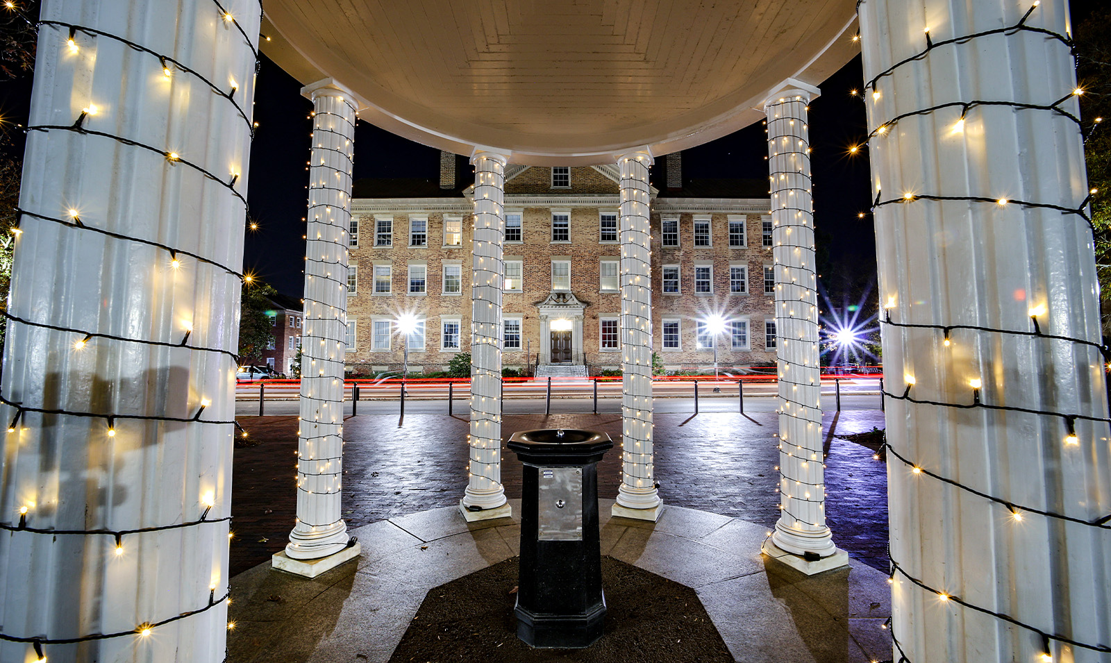 Nightime view from inside the Old Well on the campus of UNC-Chapel Hill. Pictured is the well's black fountain. Holiday lights are seen on the structure's white beams, and in the background are passing cars on Cameron Avenue and South Building.