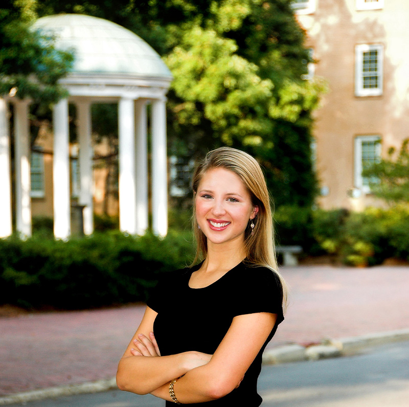 A portrait of former UNC-Chapel Hill Student Body President Eve Carson. She is standing on a brick pathway and in the background, on the other side of Cameron Avenue, is the Old Well.