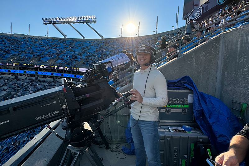 A man, Cam Neuwirth, operating a large broadcast camera while working at a Carolina Panthers game at Bank of America Stadium.