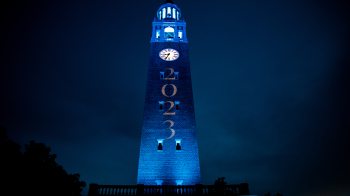 The Morehead-Patterson Bell Tower lit up in Carolina Blue at night with highlighted white text reading 