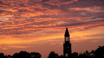 Morehead-Patterson Bell Tower on the campus of UNC-Chapel Hill at sunset with clouds weaving in and out of an orange and pink sky on a summer evening.