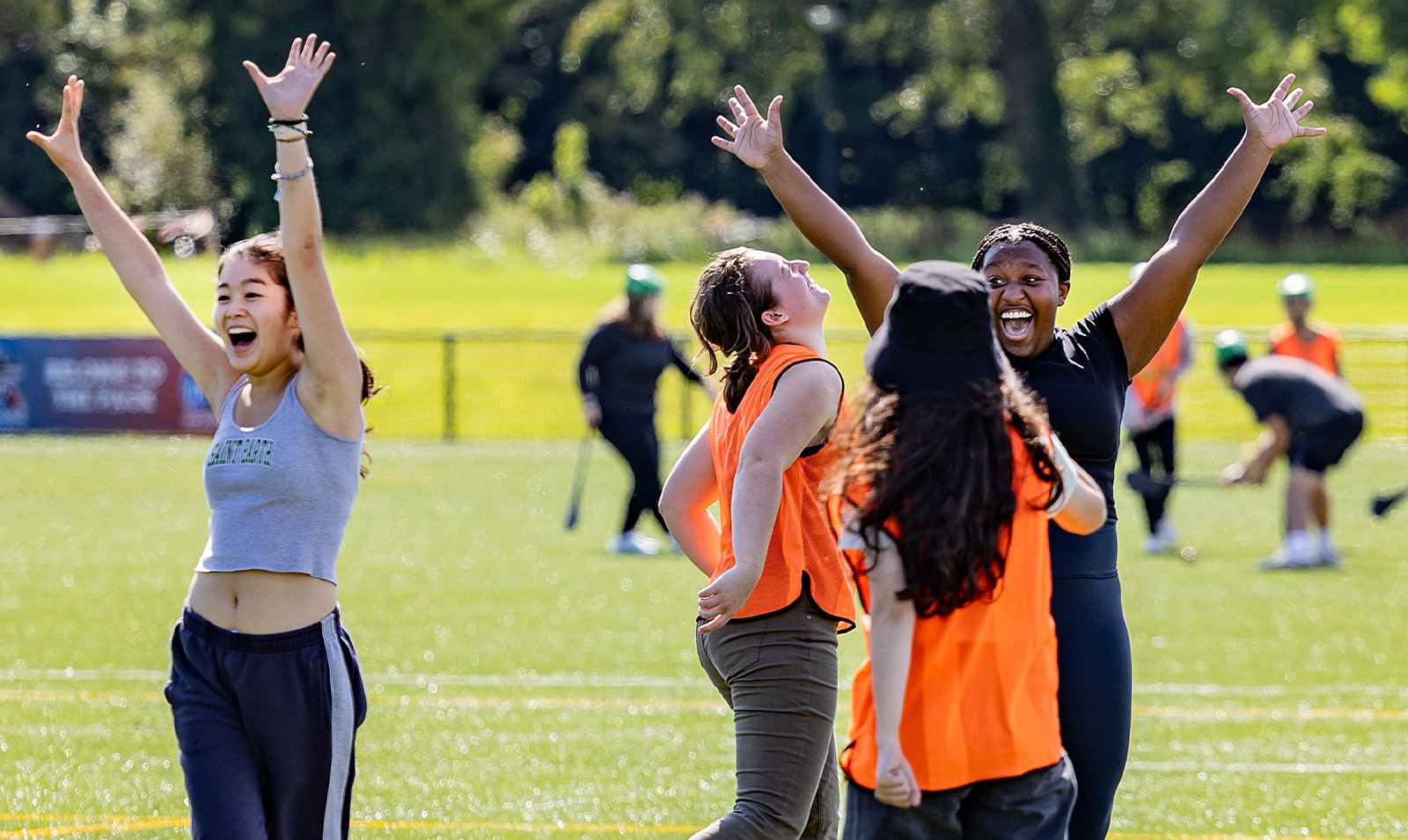 Students, some wearing orange pinnies, raising their arms in celebration while doing a group sport activity on a turf field.