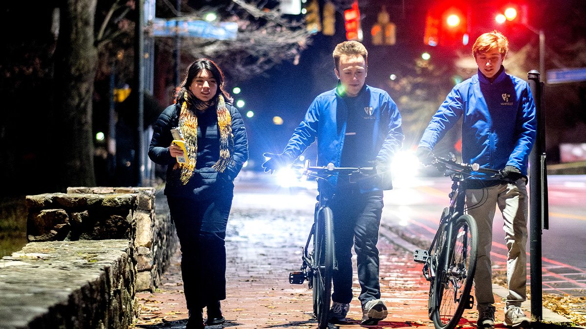 A woman and two men with bikes walking on a brick pathway on the campus of UNC-Chapel Hill at night.