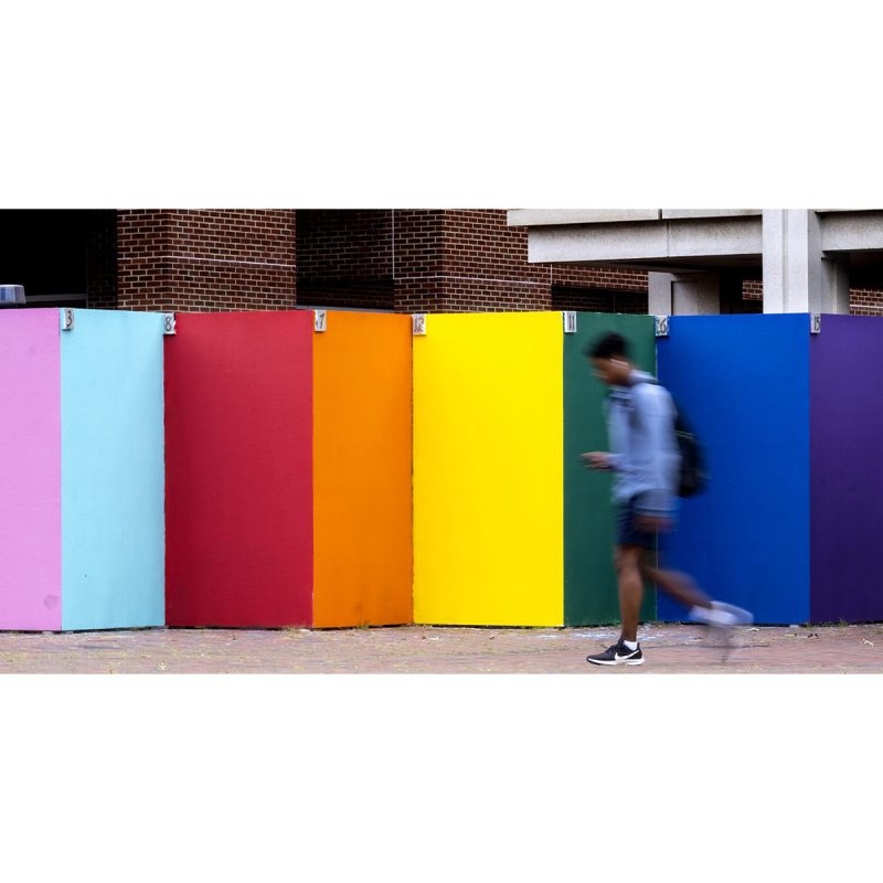 A student walking in front of mural boards painted colors of the rainbow.