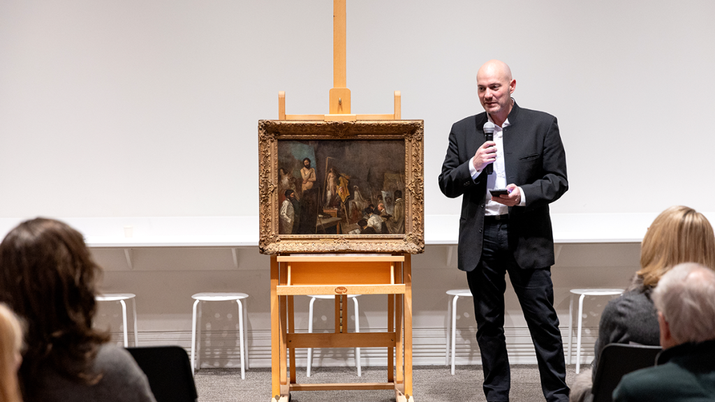 Raphael Falk stands next to painting wearing suit and speaking to audience while reading his speech from iPhone. 