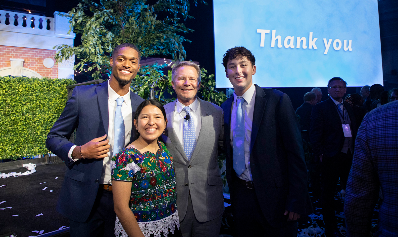 Chancellor Kevin M. Guskiewicz posing with three students at a fundraiser celebration event. In the background on a projector screen is a slide reading 