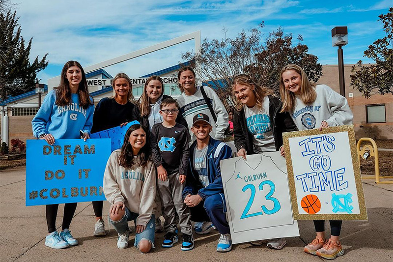 A group of eight college students posing for a photo with 9-year-old Colburn Dean in front of his school. The students are holding up signs that read "Dream It, Do It #Colburn," a No. 23 Colburn basketball jersey and "It's Go Time".