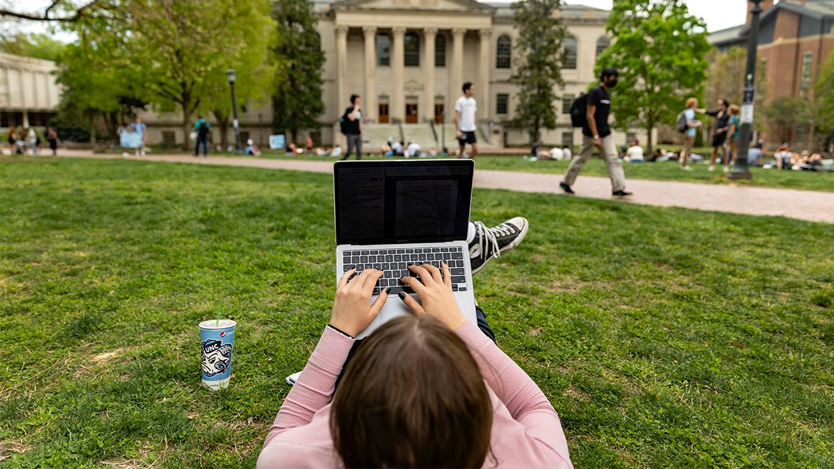 Student laying down on the main quad with a laptop placed on their legs.