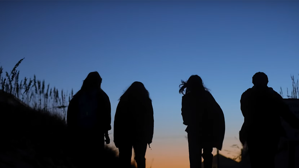 Four individuals walking on the beach during sunset, with their silhouetted backs to the camera.