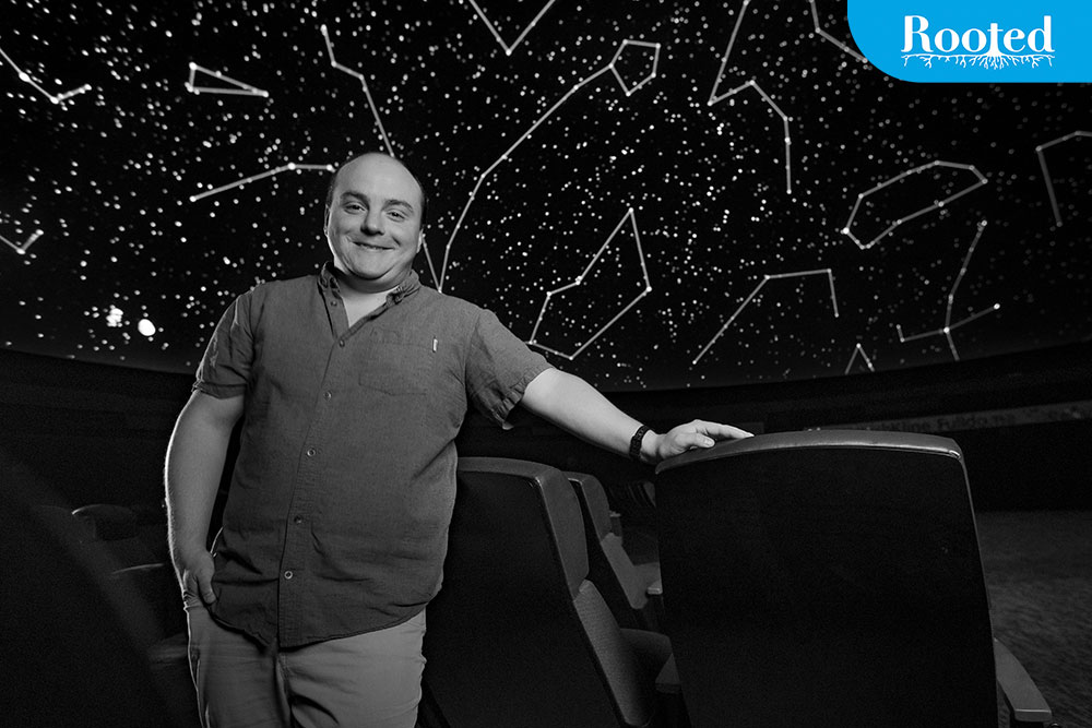 Black and white photo of Nick Eakes wearing a button-up and standing in front of the planetarium's massive screen.