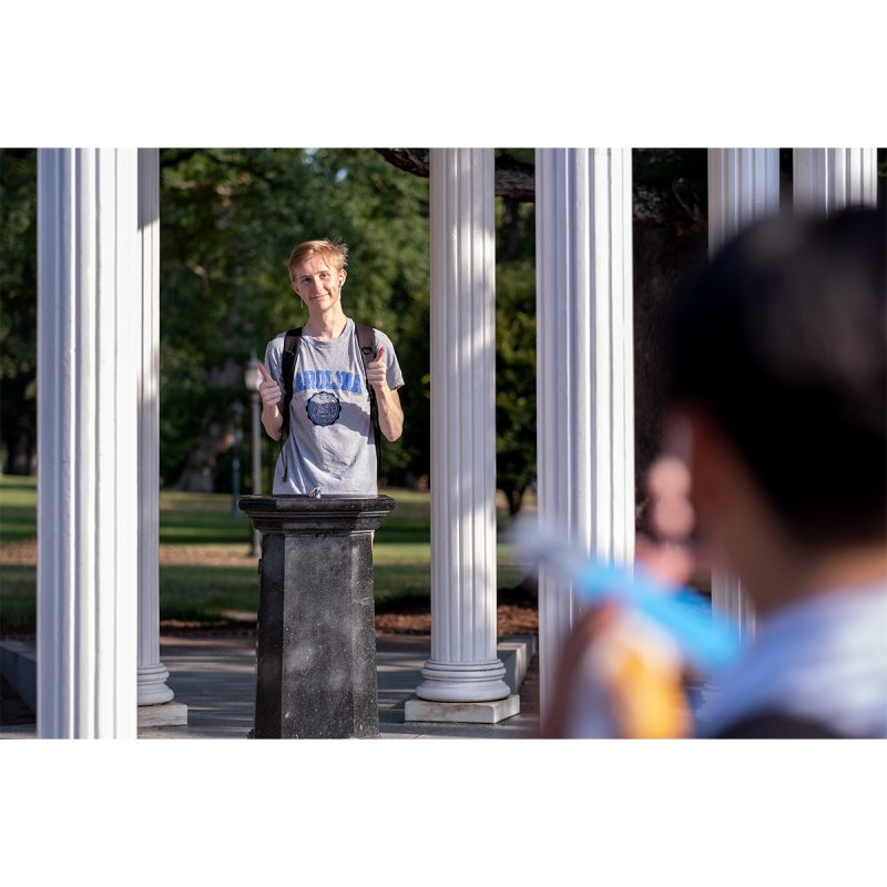 A student smiling and giving double thumbs up while posing for a photo behind the fountain of the Old Well on the campus of UNC-Chapel Hill.