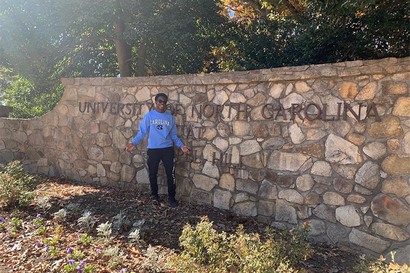 Rotimi Kukoyi standing in front of a stone sign on the campus of UNC-Chapel Hill reading "University of North Carolina at Chapel Hill".