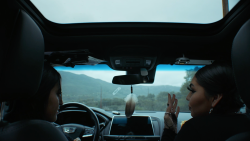 Two women speaking in the front seat of a car with a mountain range outside of their front window.