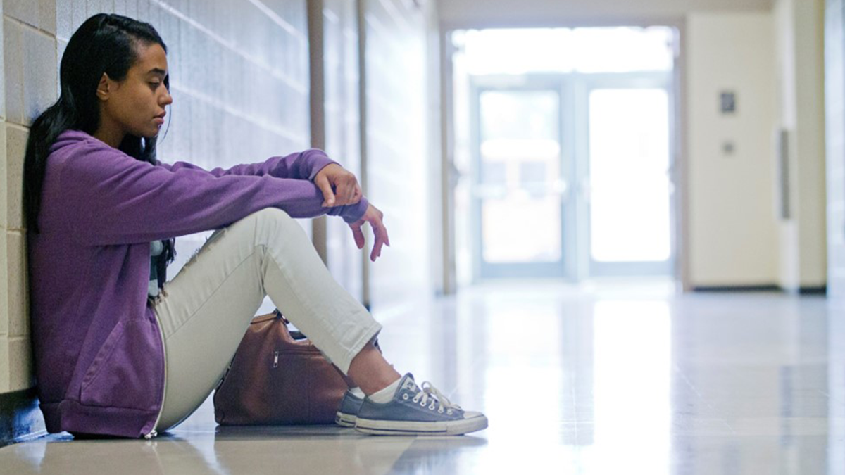 Young girl sitting against white brick wall of a school hallway.