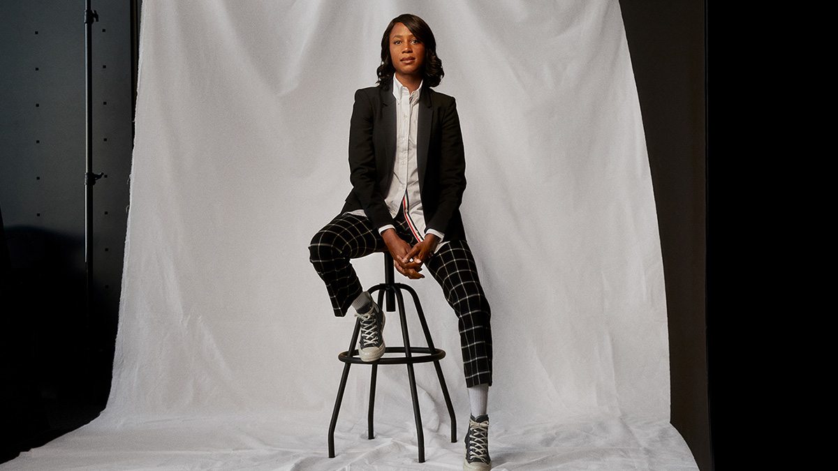 Ilana Finley sitting in front of white backdrop with black suit jacket, white shirt, patterned pants and converse shoes.