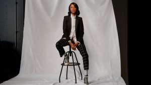 Ilana Finley sitting in front of white backdrop with black suit jacket, white shirt, patterned pants and converse shoes.