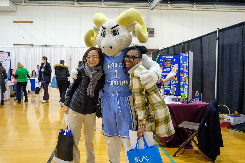 Two faculty staff members smiling and standing next to Rameses the Ram with his arms around them.