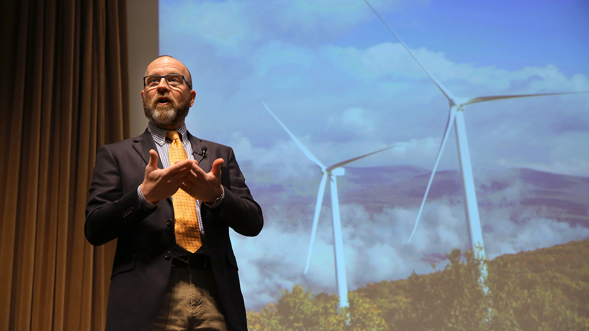 Man wearing microphone and speaking in front of powerpoint image with windmills.