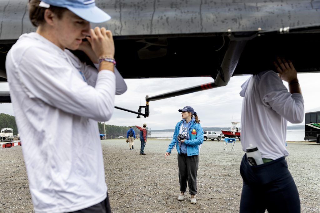 Karen Jordan standing on beach wearing a blue jacket and baseball cap as she watches the rowing team prepare.