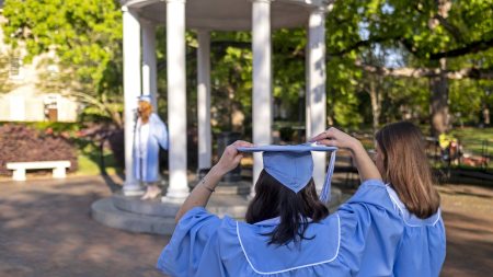 A student wearing a cap and gown with her back to the camera. She's waiting by the Old Well as another student in regalia has a graduation photo taken at the Old Well.