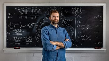 A graduate student, Andrew Adair, posing for a portrait with his arms crossed in a classroom. Behind him is a black chalkboard full of math equations written with white chalk.