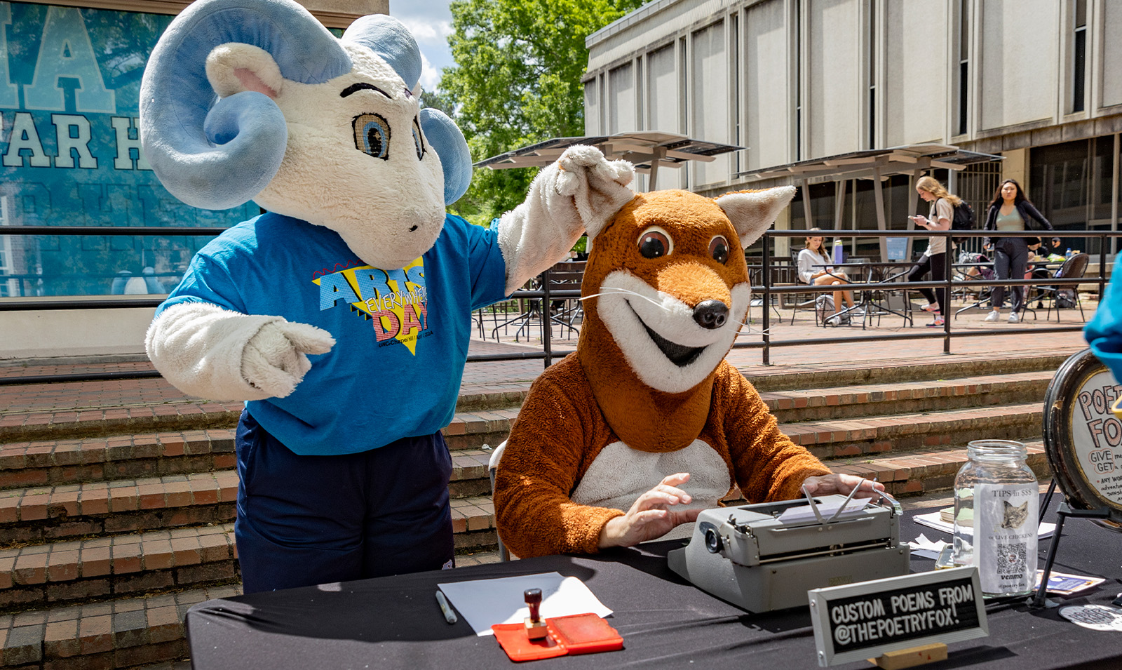 A ram mascot, Rameses Jr., holding the ear of another mascot, Poetry Fox, at Arts Everywhere Day.