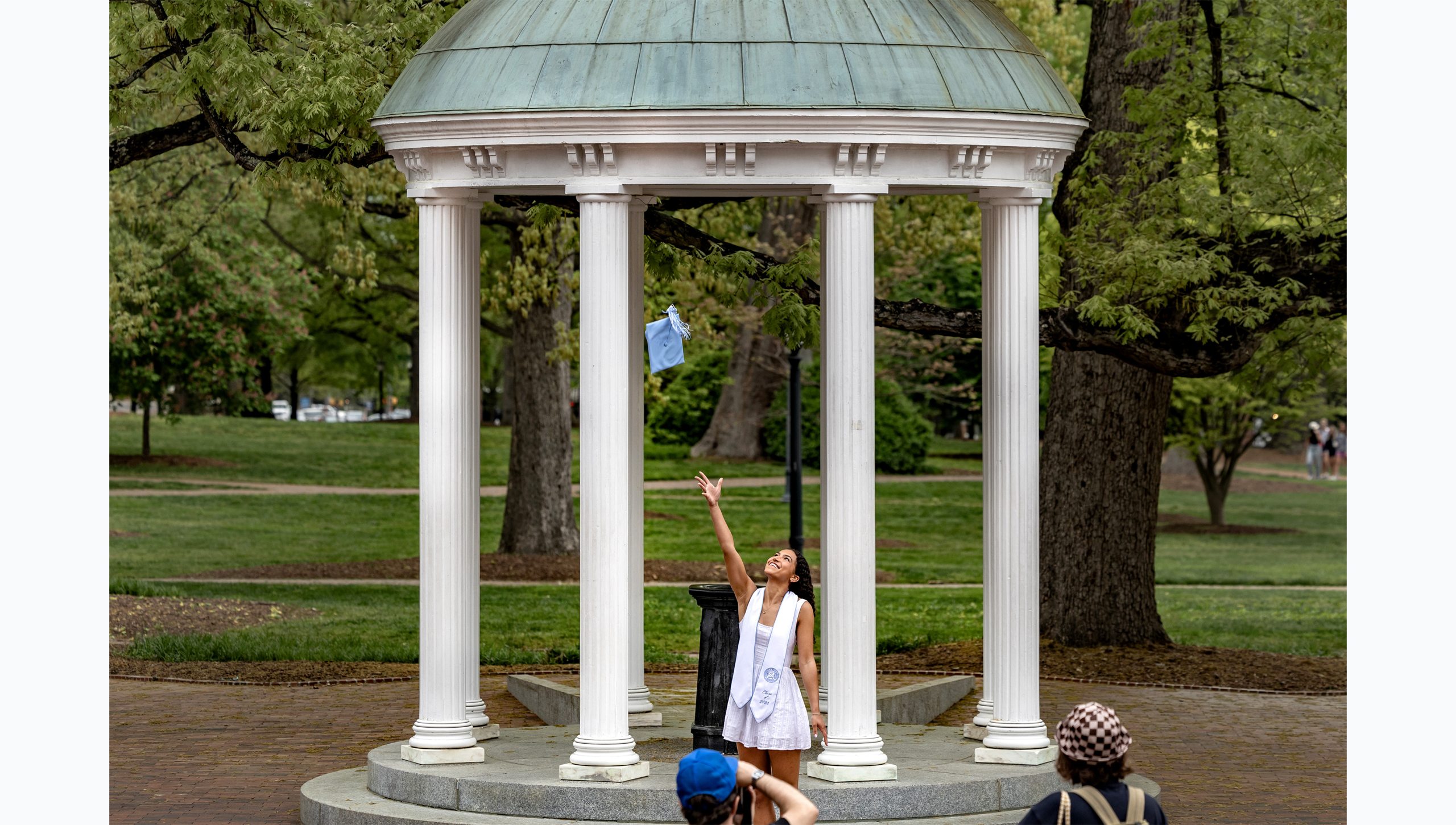 A student tossing her cap up in the air as she poses for a graduation photo at the Old Well.