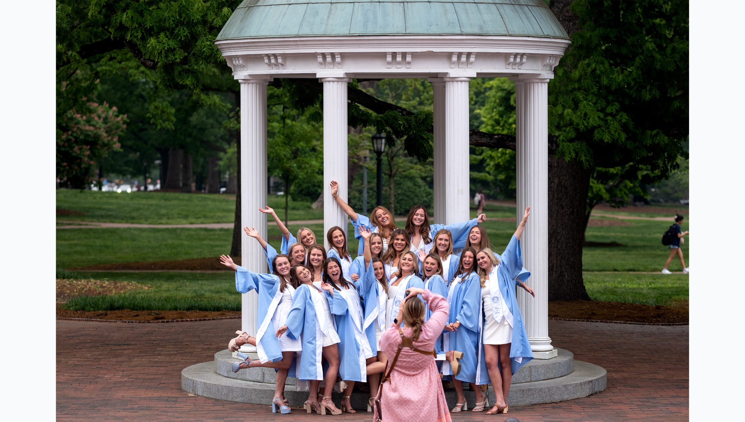 Group of students throw hands in air while posing in graduation gowns in front of Old Well