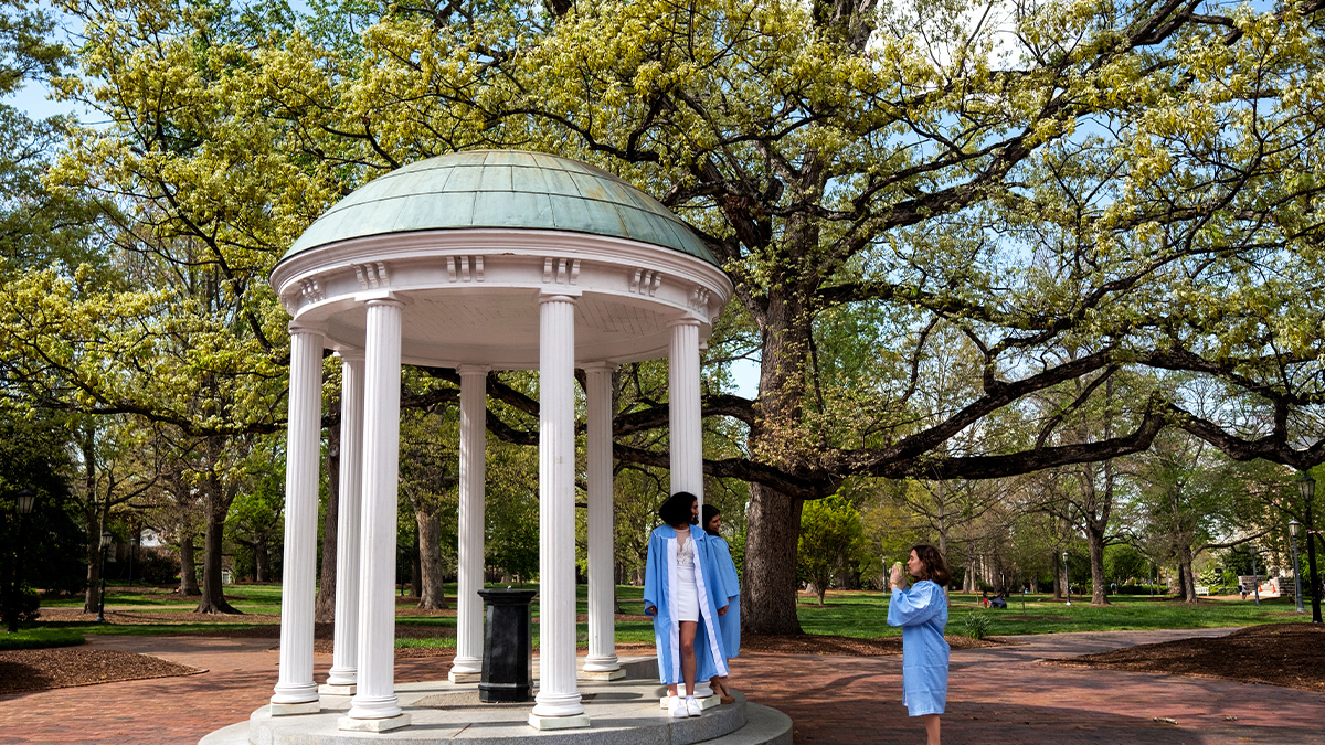 A student in a cap and gown taking an iPhone camera of two other students in caps and gowns posing by the Old Well on the campus of UNC-Chapel Hill