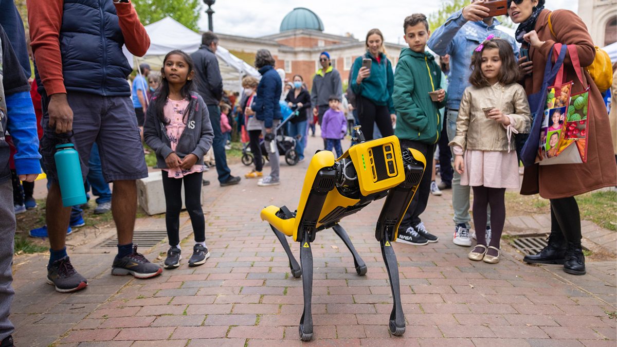 Boston Dynamics four legged robot walking around campus with onlookers of all ages surrounding it.