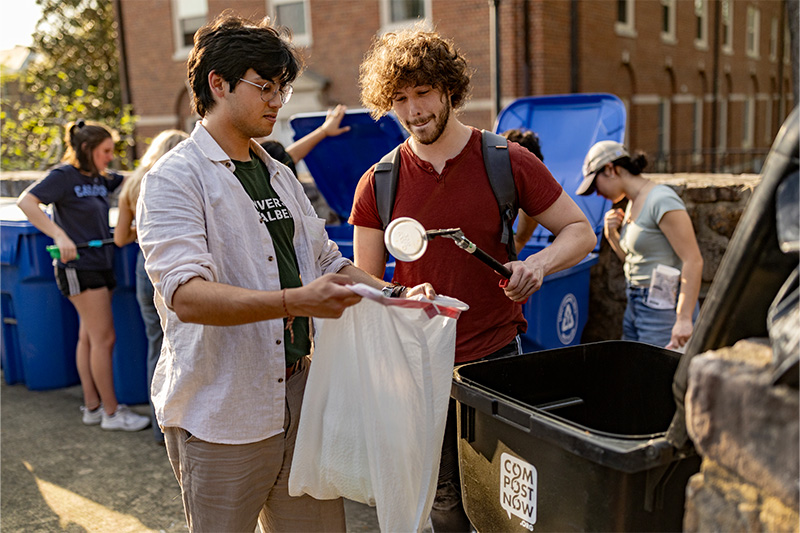 Two students, one holding a trash bag and the other placing a plastic lid inside of it.