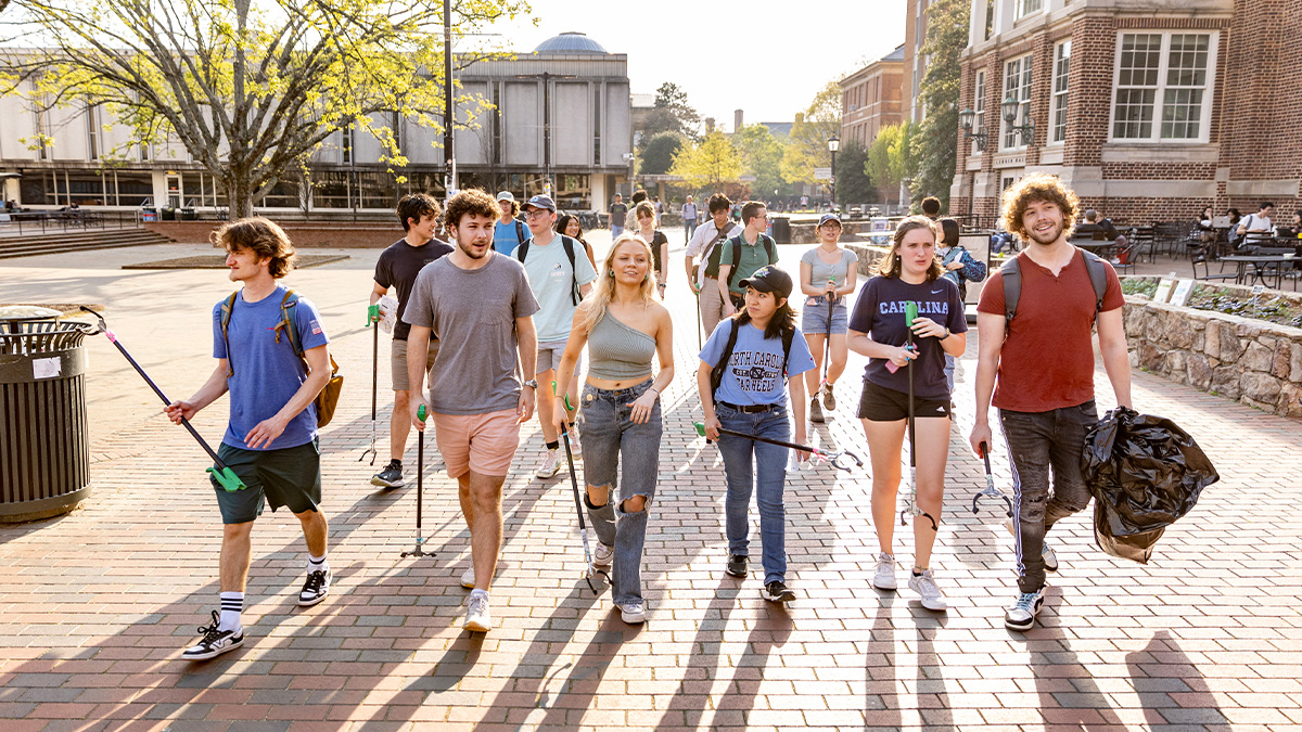 Students walking along brick pathways on the campus of UNC-Chapel Hill holding trasha pickers and trash bags.