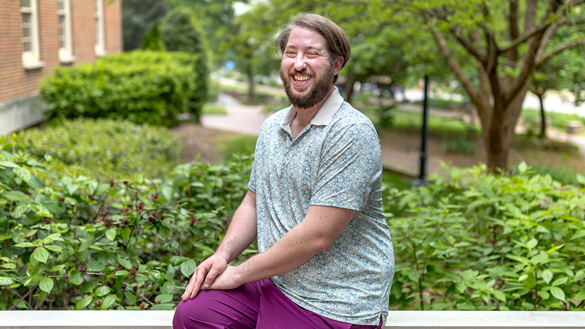 Mark Radin sitting in front of campus scenery wearing green button up and purple pants.