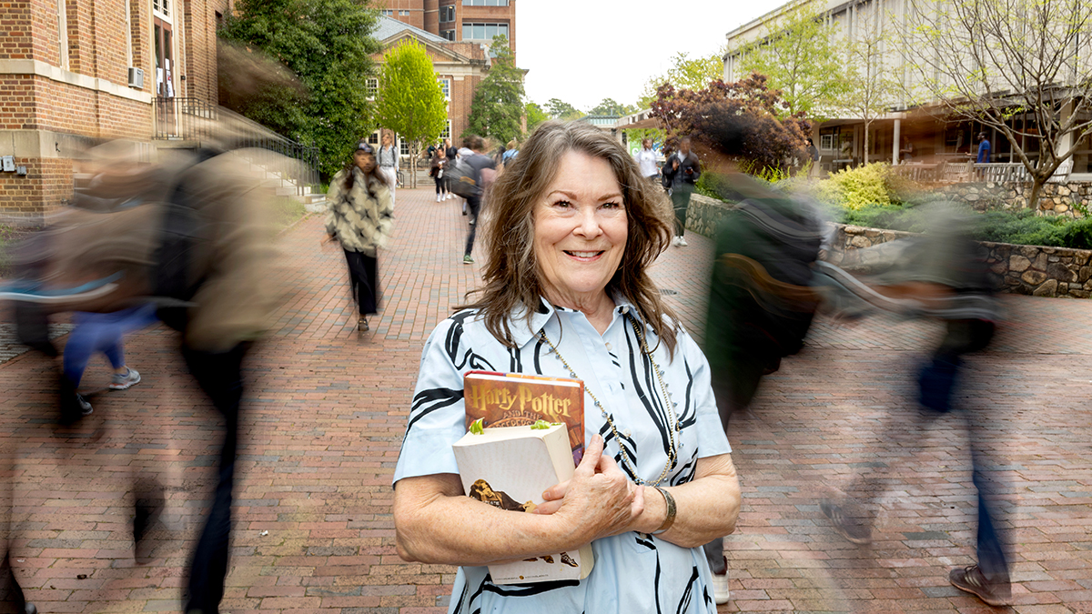 Janet Vitolo standing on campus with book in her hand while fellow students walk around her.