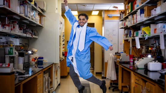 Rami Darawsheh jumping in lab room while wearing Commencement robes.